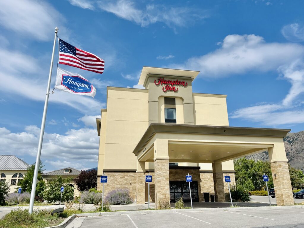 [Picture of the front entrance of a Hampton Inn hotel, clear skies, American flag.]